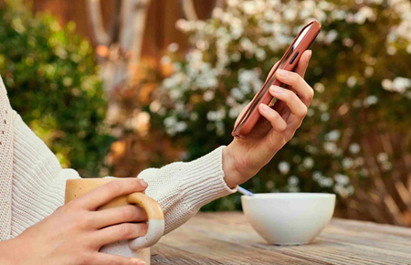 Person hält ein Smartphone in der Hand und trinkt Kaffee im Garten; Fokus auf mobile Nutzung im Alltag.